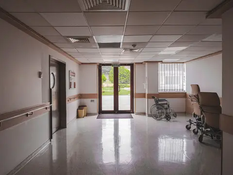 An empty hospital corridor with an unoccupied wheelchair and a stretcher chair by glass doors that open to a garden