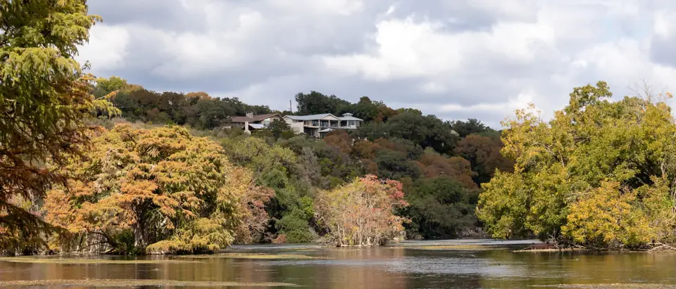 A Texas lake framed by cypress and oak trees in autumn colour, with houses on the far hill near New Braunfels